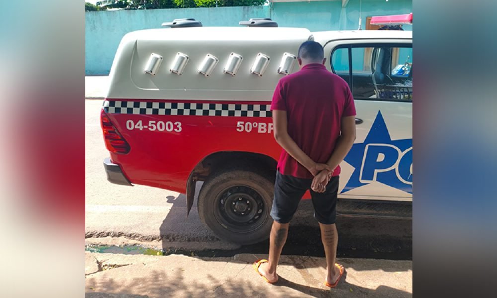 Homem de costas com mãos para trás ao lado de uma viatura policial.