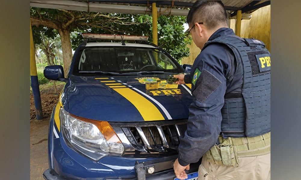 Policial da PRF ao lado de viatura azul com emblema no capô.