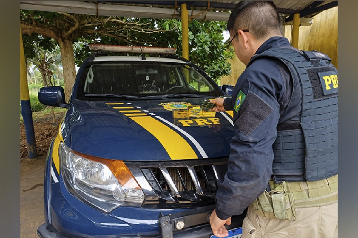 Policial da PRF ao lado de viatura azul com emblema no capô.