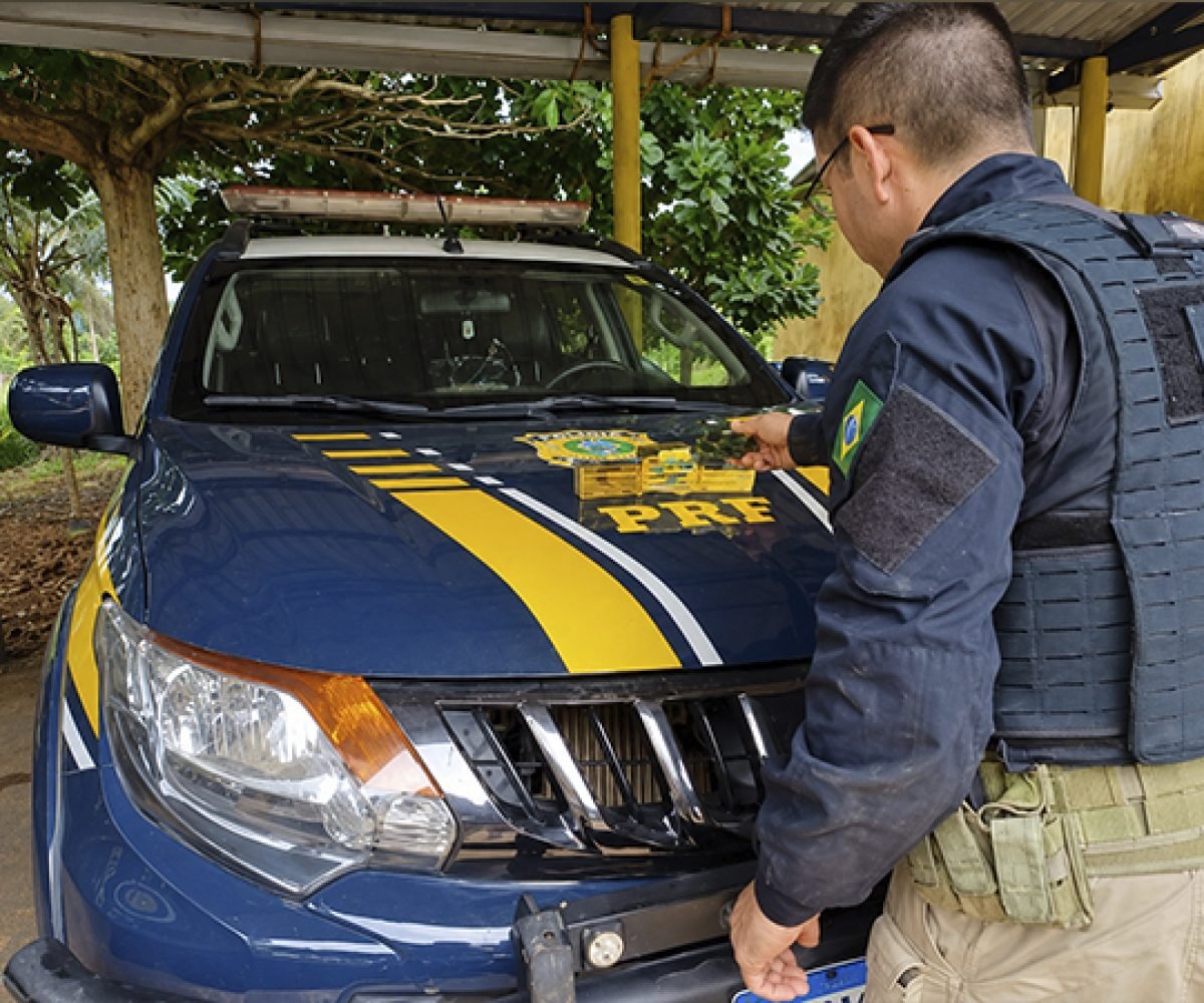 Policial da PRF ao lado de viatura azul com emblema no capô.
