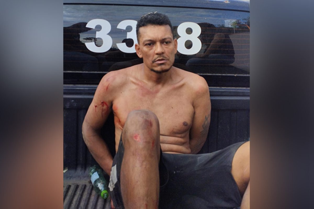 Homem sem camisa com ferimentos e algemado em parte traseira de veículo policial.