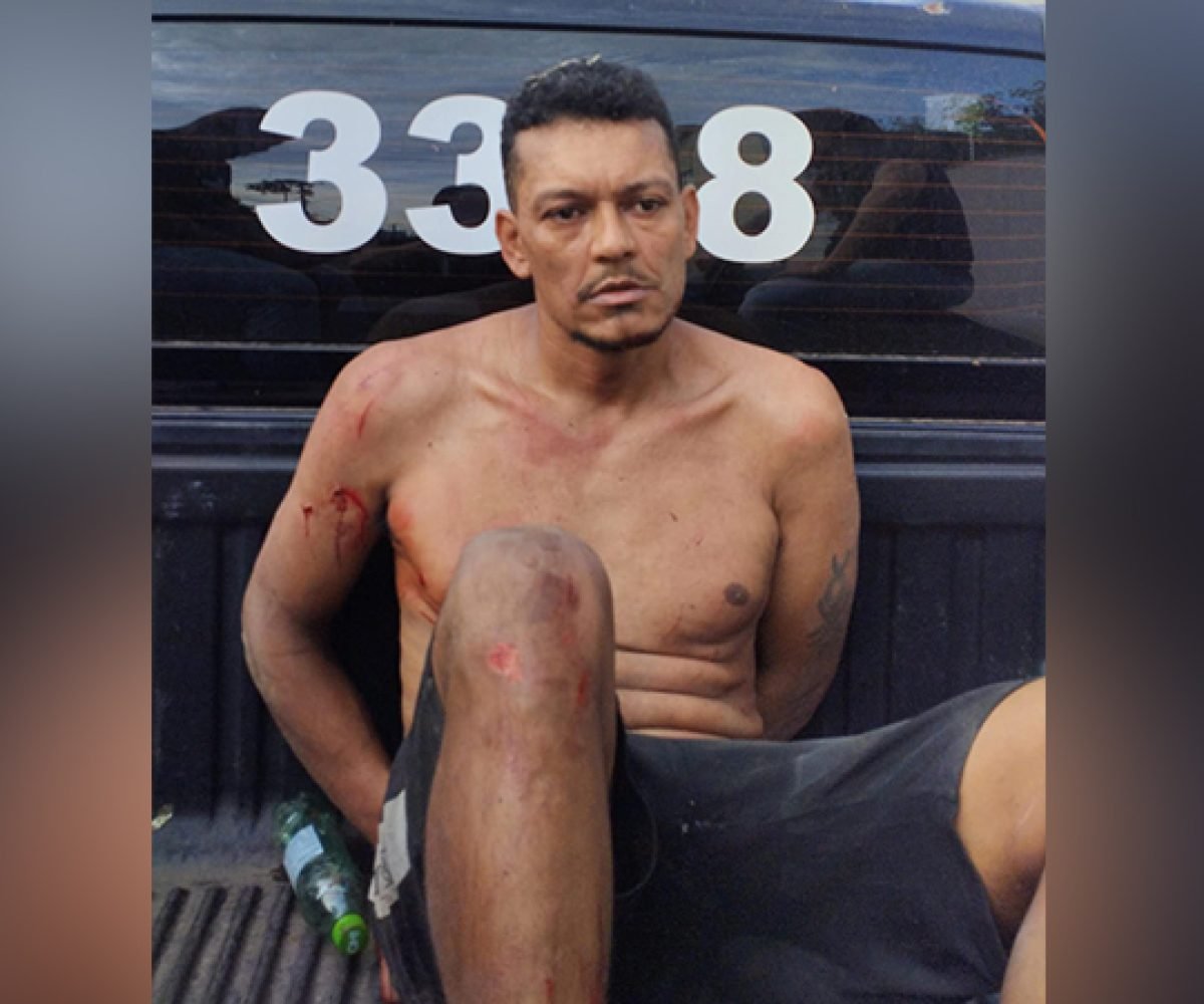 Homem sem camisa com ferimentos e algemado em parte traseira de veículo policial.