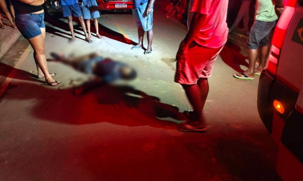 Corpo desfocado de uma pessoa caída na rua, cercado por curiosos e luzes vermelhas de emergência.