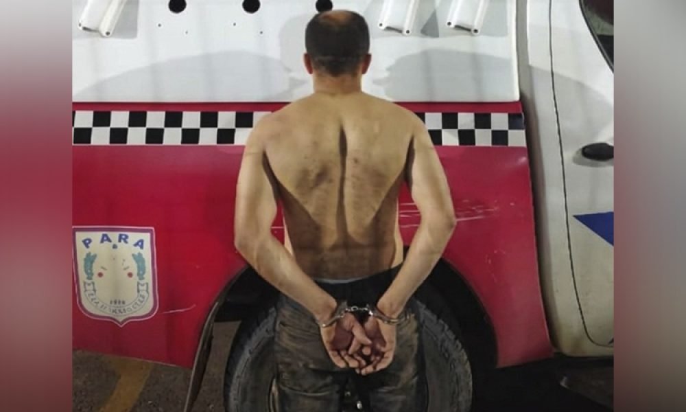 Homem algemado de costas, sem camisa, em frente a veículo policial.