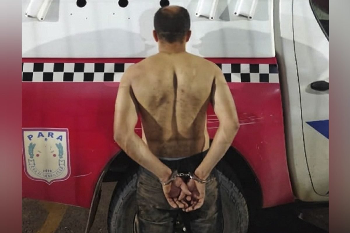 Homem algemado de costas, sem camisa, em frente a veículo policial.