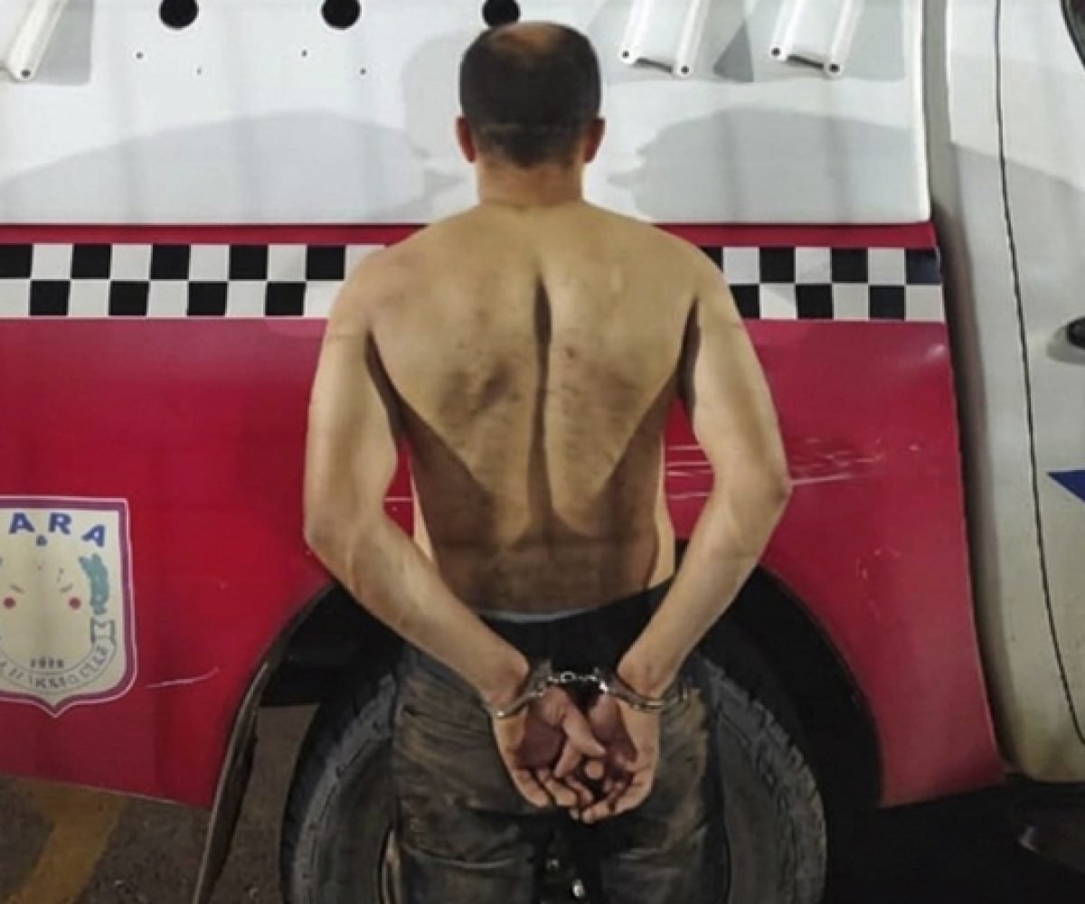 Homem algemado de costas, sem camisa, em frente a veículo policial.