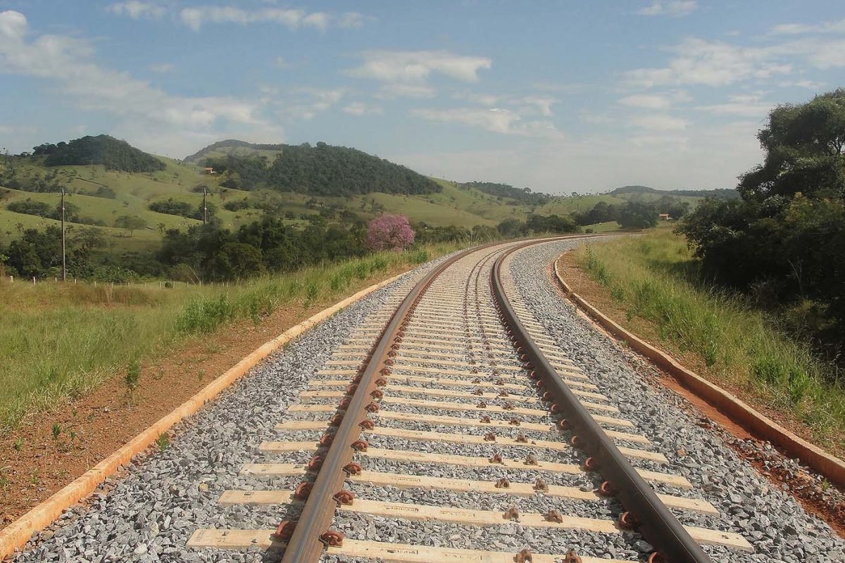 Trilhos de trem em curva atravessando uma paisagem rural montanhosa em dia ensolarado